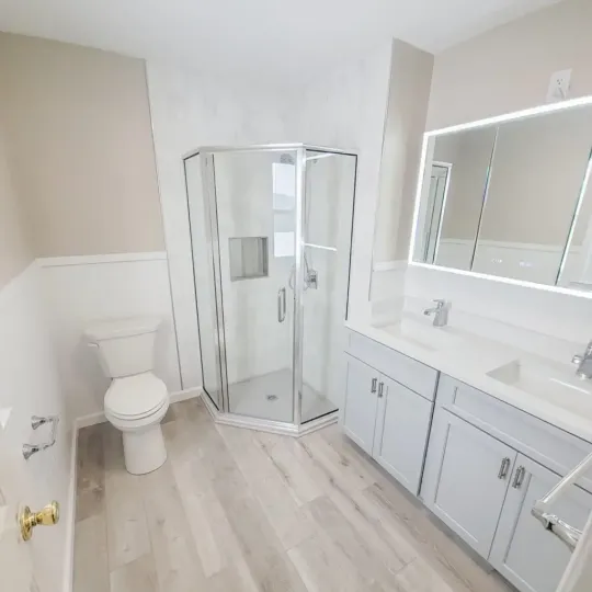 Bathroom remodel with corner glass shower, light gray double vanity, and backlit LED mirror.