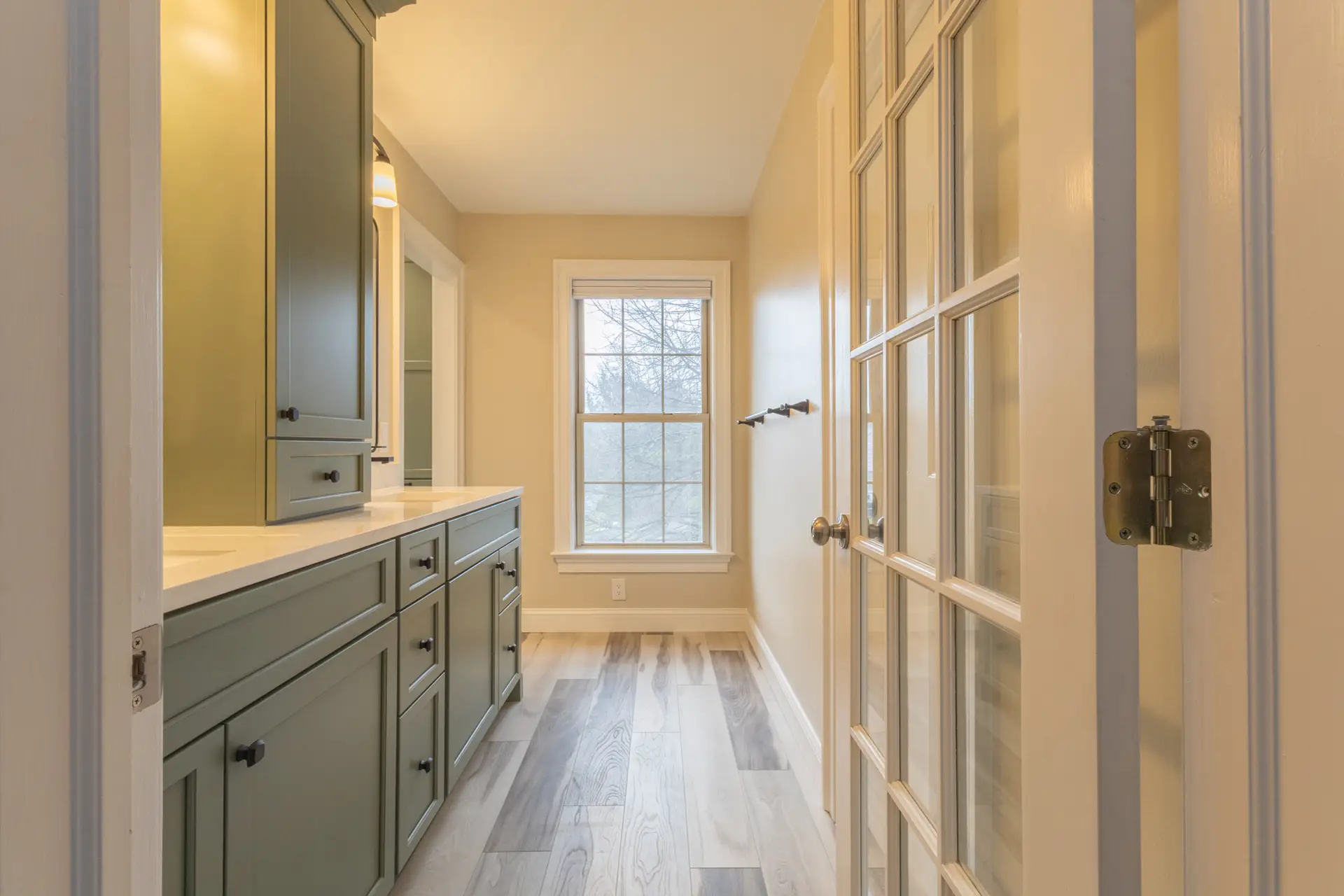 Doorway opening into bathroom entryway with double-sink vanity, sage green cabinets and counter-to-ceiling cabinet between the sinks, and a window at the end of the room