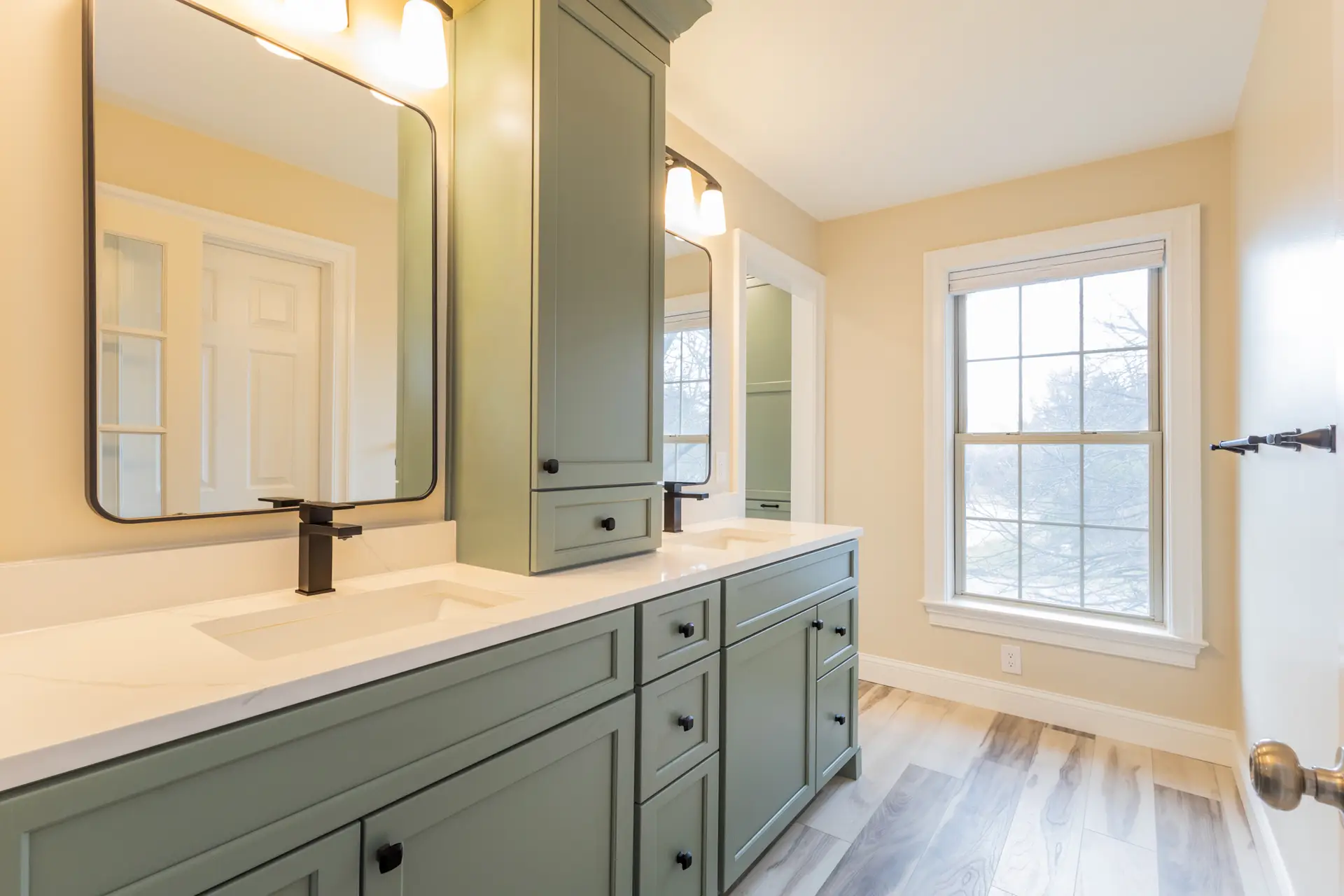 Sage green double-sink vanity with matte black fixtures and bright, warm lighting. Window at the end of room next to doorway